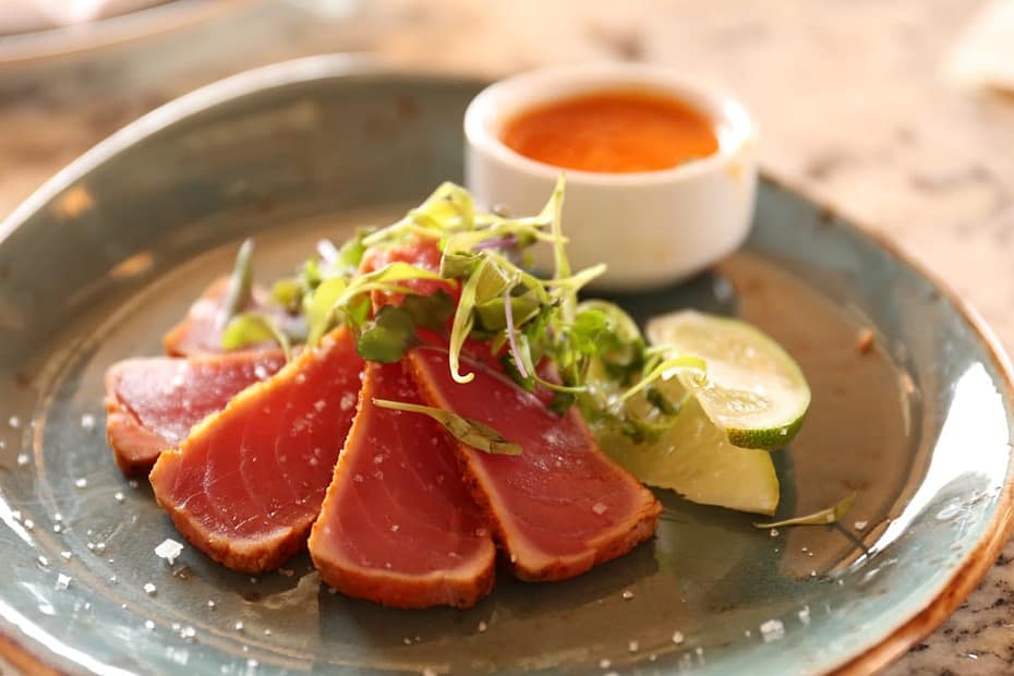 Plated rare sliced pan seared ahi tuna steaks with ruby red centers, sesame crust, microgreens, lime wedge, and dipping sauce