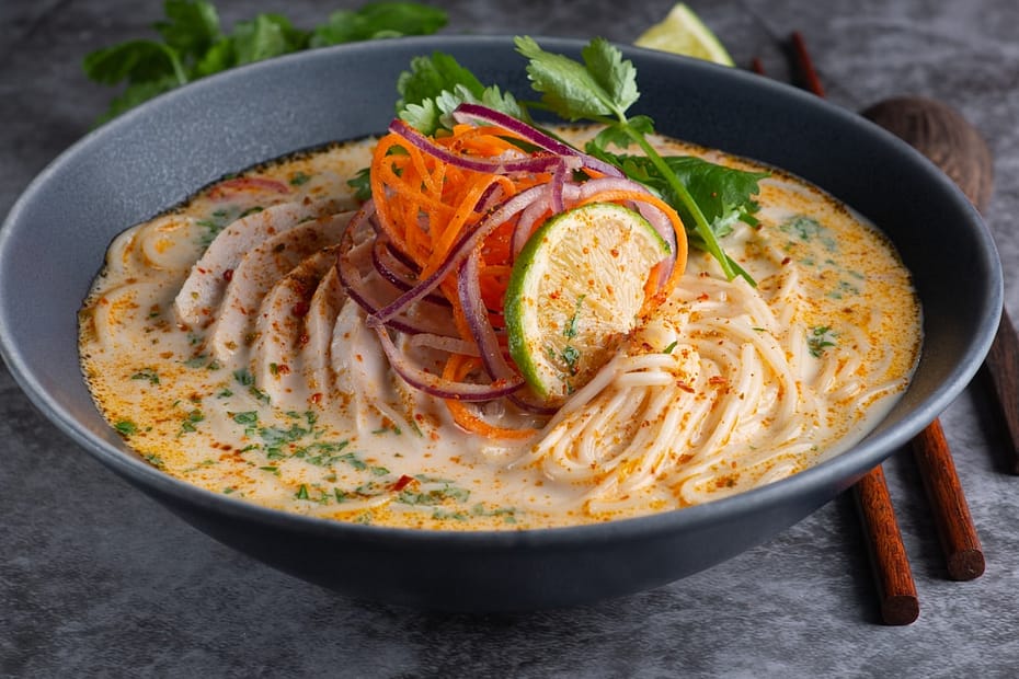 Steaming bowl of creamy Thai coconut curry chicken soup with tender sliced chicken, vermicelli noodles, fresh lime wedge, and aromatic herbs